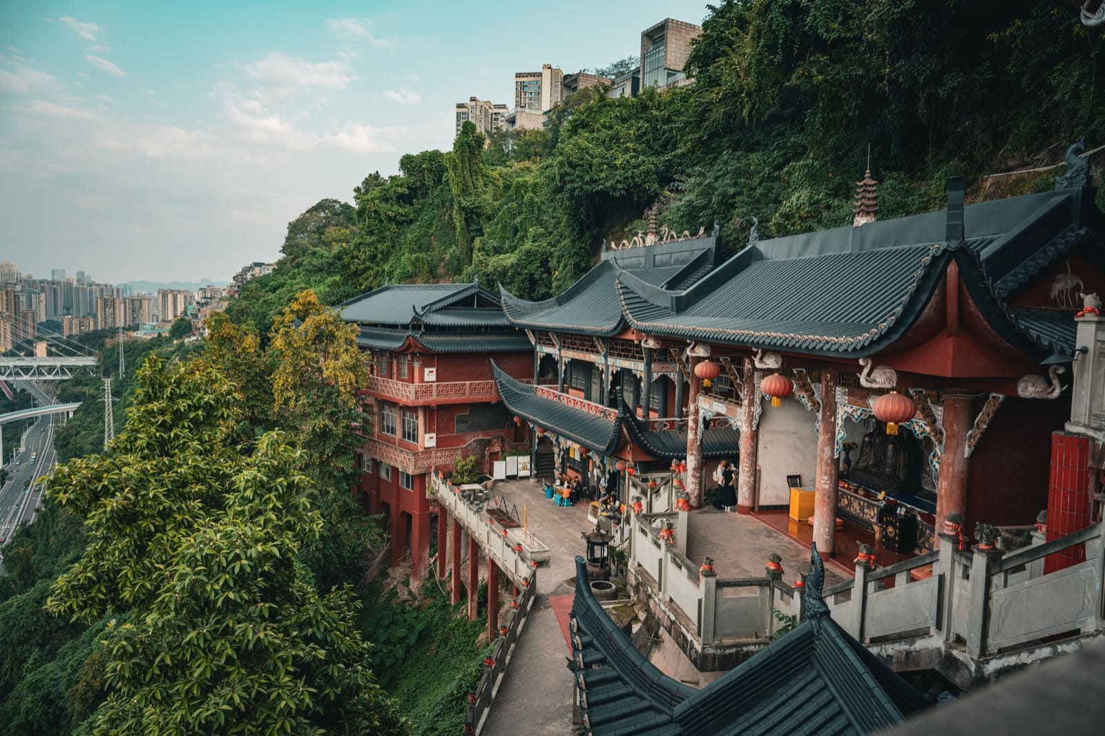 A traditional Chinese temple complex built into a hillside, featuring dark tiled roofs and red pillars, with a scenic view of a modern highway bridge and urban skyline in the distance.