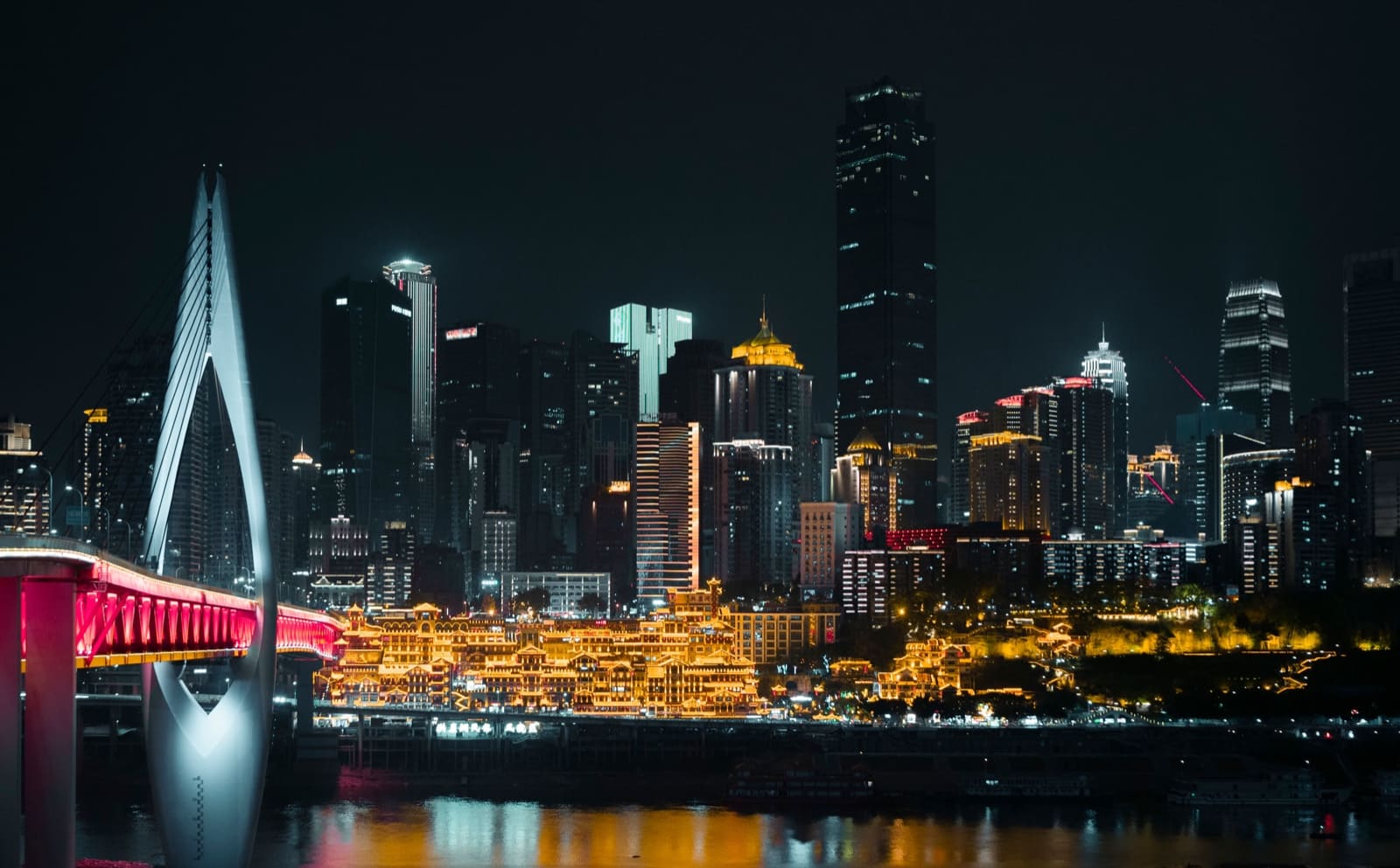 A vibrant night view of a modern city skyline featuring illuminated skyscrapers, contrasted by a foreground of traditional-style buildings bathed in warm golden light, with a large, bright white cable-stayed bridge spanning across a dark river.