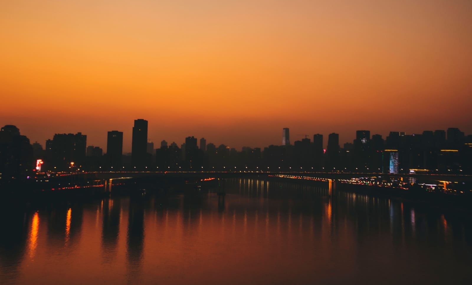 A silhouetted cityscape at sunset with an orange sky reflecting over a calm river, featuring a bridge illuminated with small lights crossing the water.