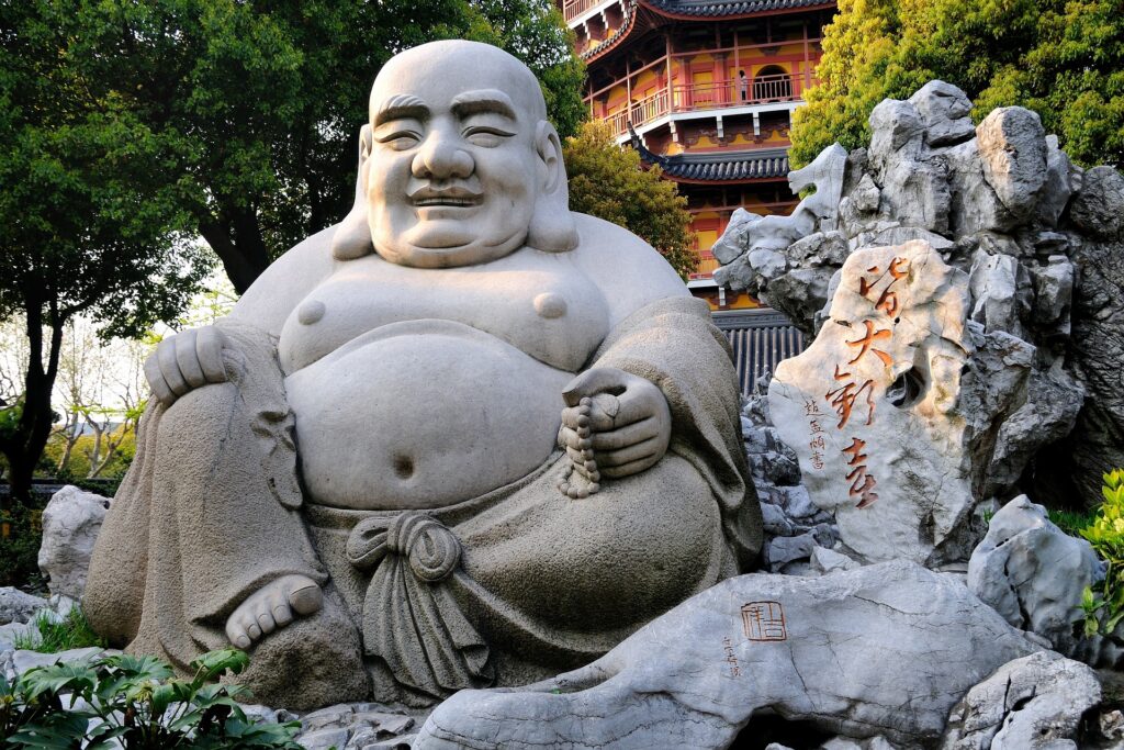 A large, stone statue of a joyful Laughing Buddha sitting in a serene garden, accompanied by a textured rock formation engraved with Chinese calligraphy and a traditional multi-story pagoda in the background.