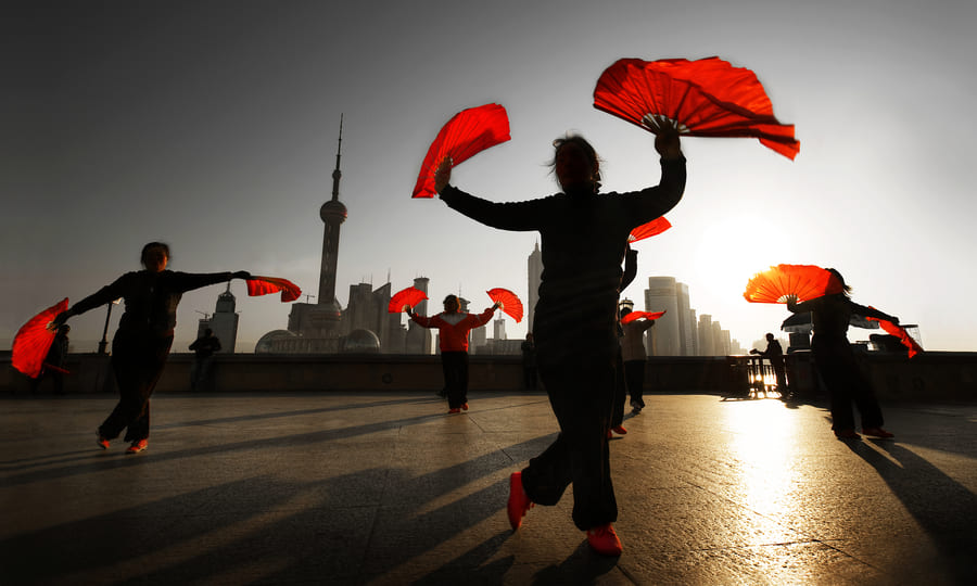 A group of people performing a traditional Chinese fan dance outdoors at sunrise, silhouetted against the iconic Shanghai skyline and the Oriental Pearl Tower.