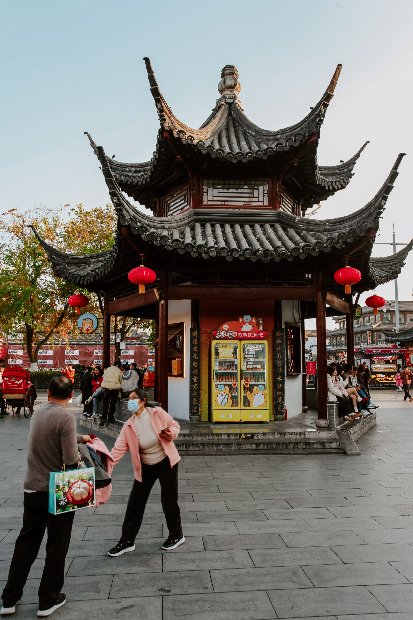 A traditional Chinese pavilion with curved, tiled roofs and hanging red lanterns, serving as a kiosk featuring a bright yellow vending machine, situated in a paved public square with pedestrians passing by.