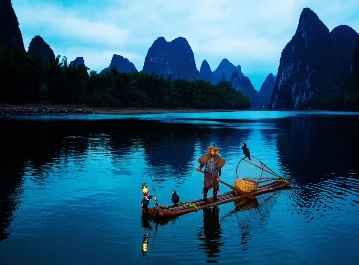 A traditional fisherman wearing a conical hat and cape stands on a bamboo raft on a calm, blue-toned river, accompanied by two cormorant birds and a lantern, set against a backdrop of dramatic karst mountains.