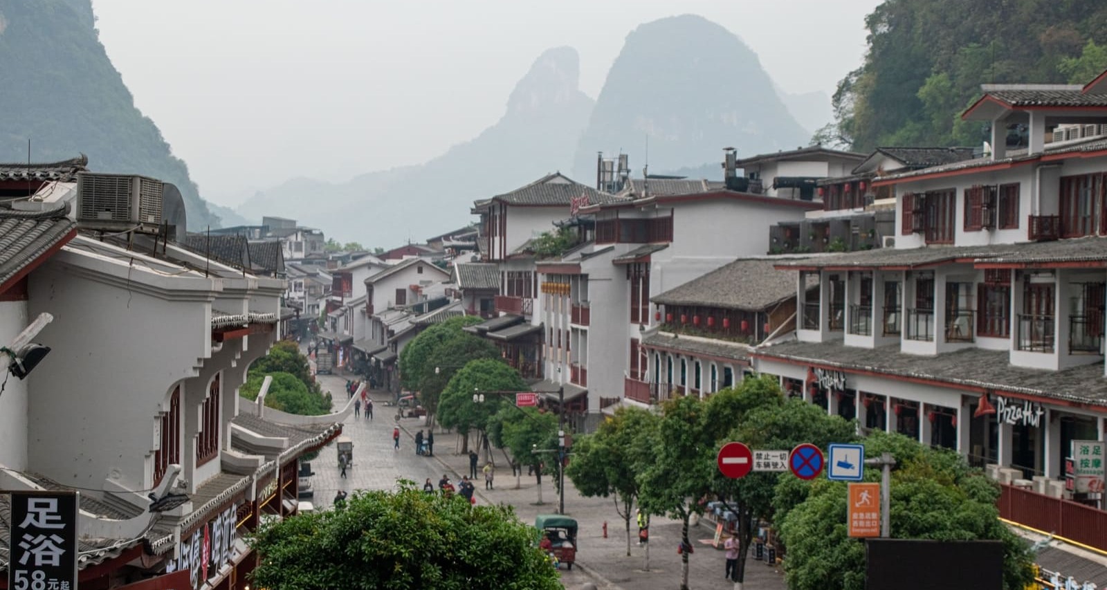 A view down a paved street in a town with traditional Chinese architecture, featuring white buildings with dark tiled roofs and red accents, leading towards distant, misty karst mountains.