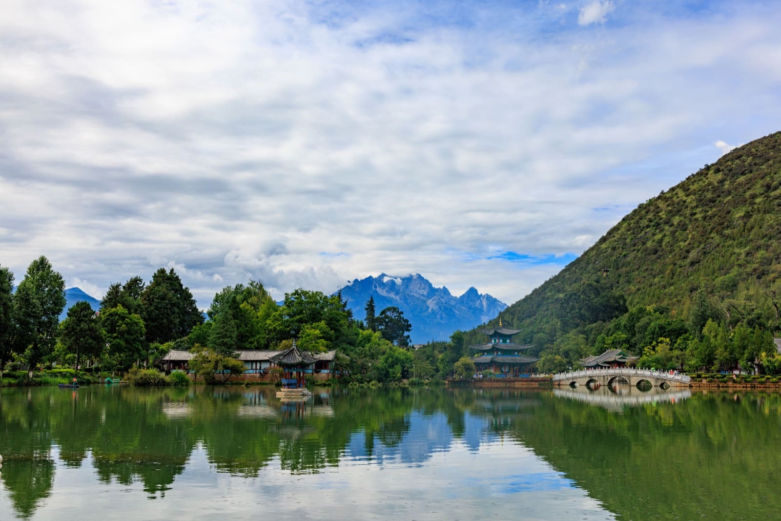Traditional Chinese pavilions and a curved stone bridge reflected in a calm lake at the Black Dragon Pool in Lijiang, Yunnan, with the snow-capped Jade Dragon Snow Mountain in the background under a cloudy sky