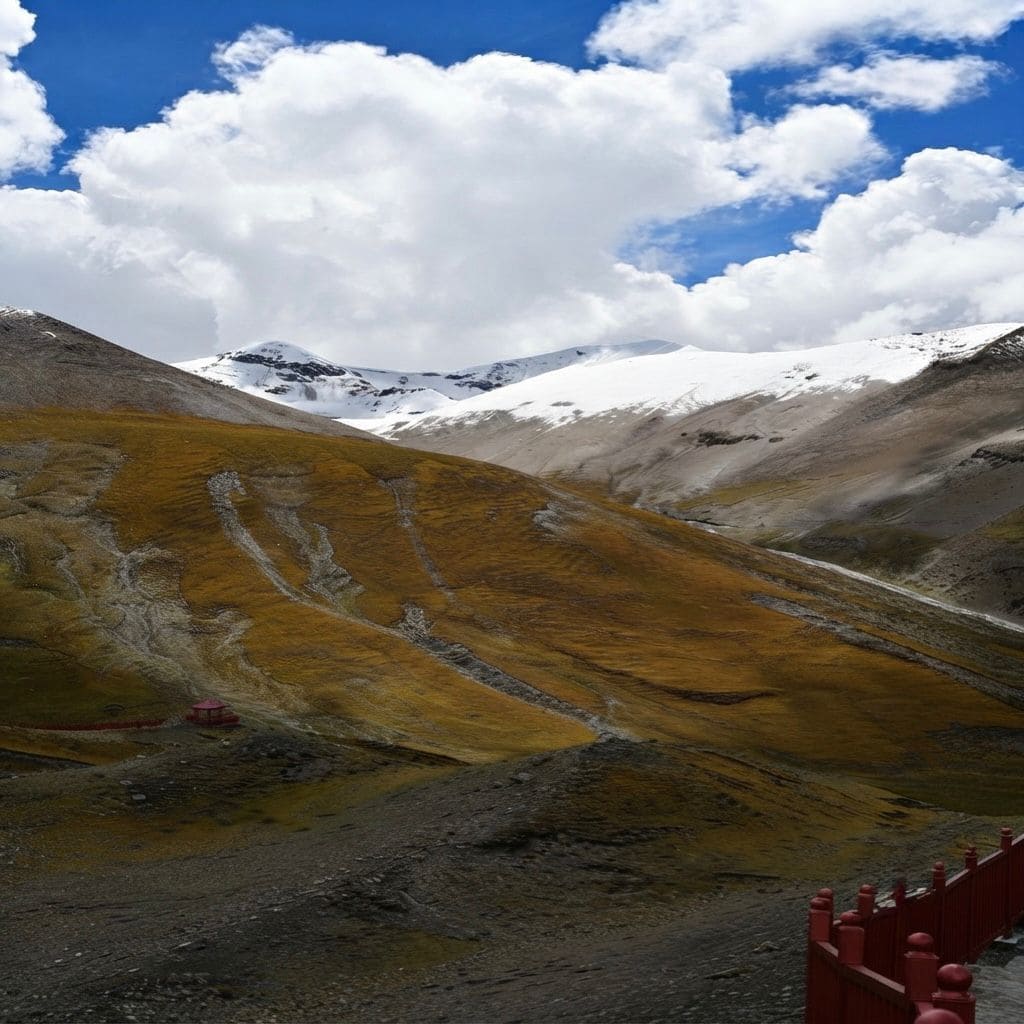 A sweeping mountain landscape featuring golden, grass-covered slopes leading up to dramatic, snow-capped peaks under a bright blue sky filled with fluffy white clouds, with a red wooden fence in the foreground.
