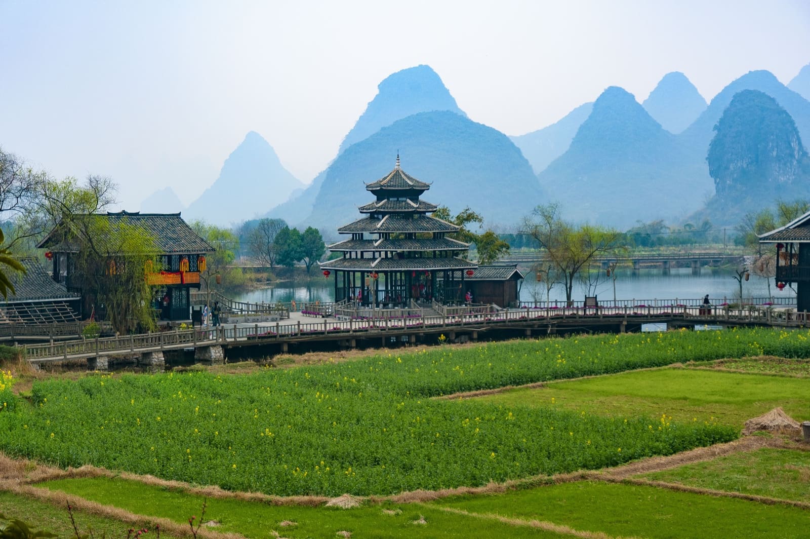 traditional, multi-tiered wooden pavilion stands along a calm river, connected by a long wooden walkway through green fields, with dramatic karst mountains rising in the misty background.